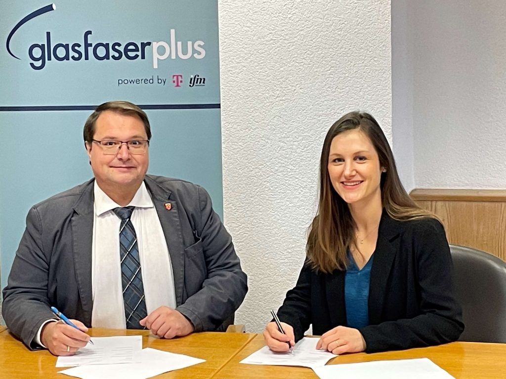 Zwei Personen sitzen an einem Tisch und unterschreiben einen Vertrag zum Glasfaserausbau in Weißdorf. Dabei lächeln sie in die Kamera.