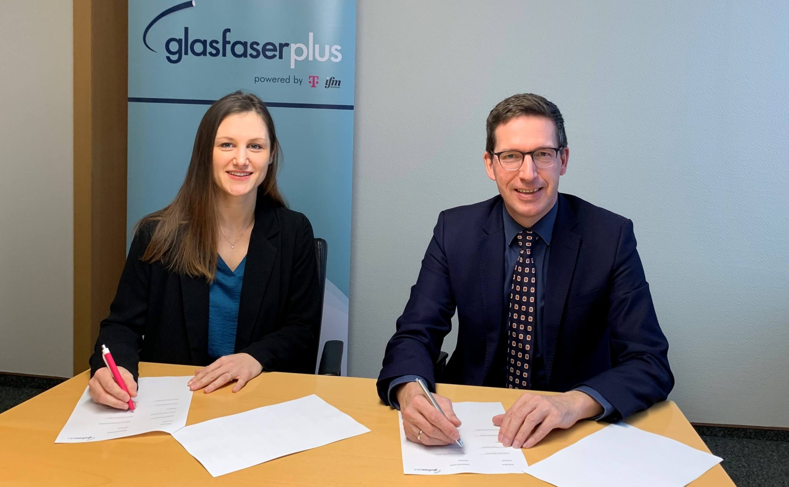 Zwei Personen sitzen an einem Tisch und unterzeichnen einen Vertrag zum Glasfaserausbau. Dabei lächeln sie in die Kamera.