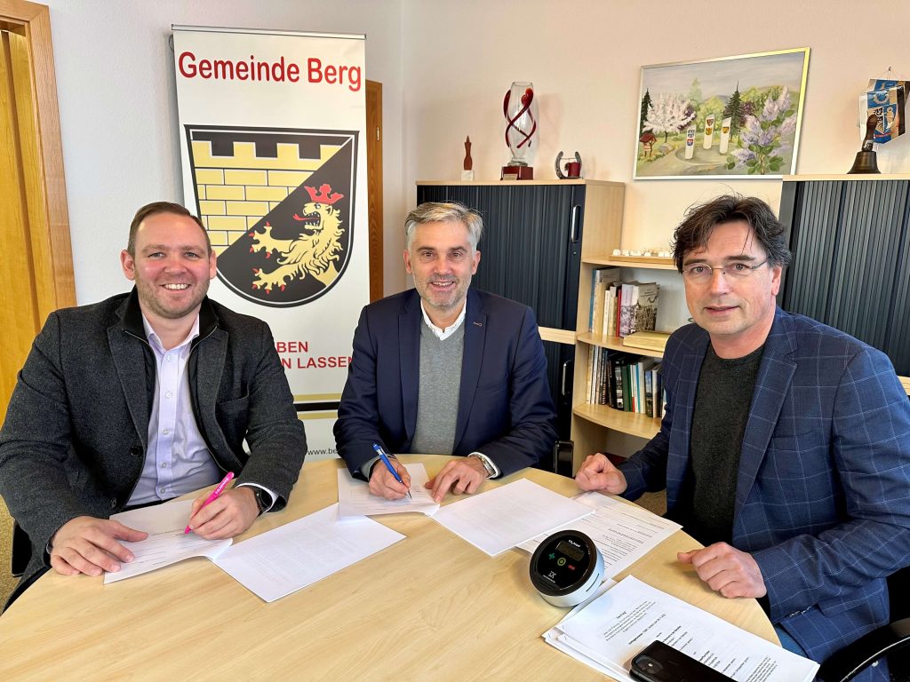 Drei Personen sitzen an einem Tisch und unterschreiben einen Vertrag zum Glasfaserausbau. Dabei lächeln sie in die Kamera.